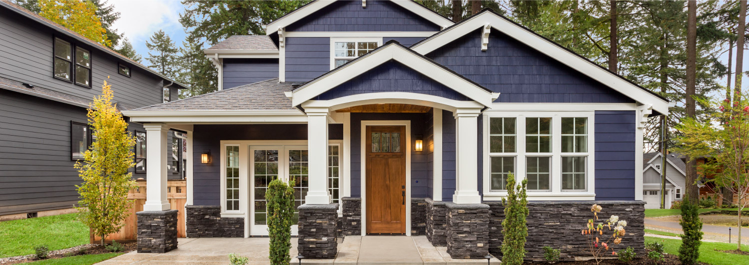 A modern suburban house with navy blue siding, white trim, and a welcoming wooden front door. The house features a covered front porch supported by white columns and surrounded by a neatly landscaped yard with small shrubs and trees.